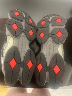 New Balance 9060 sneakers svart/grå - Säljer ett par New Balance 9060 sneakers i svart och grått med chunky sula och coola meshpaneler. Skorna har tydliga NB-loggor, snörning och detaljer i mocka och syntet. Röd detalj på plösen ger extra edge. Perfekt för dig som gillar streetwear och sportig stil.