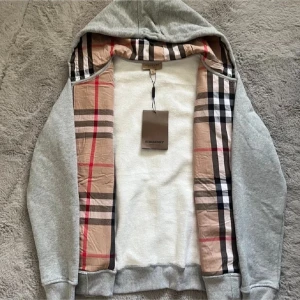Grå Burberry hoodie med rutig insida - Snygg grå hoodie från Burberry med dragkedja och huva. Insidan av luvan och öppningen har klassiskt Burberry-rutigt tyg i beige, svart och rött. Tröjan har broderad logga på bröstet, snörning i luvan och två fickor framtill. Perfekt för dig som gillar stilrena streetwear-plagg. Insidan på tröjan är grå inte vit