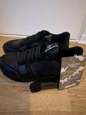 Valentino Garavani svarta sneakers - Svarta Valentino Garavani sneakers med coolt kamouflagemönster och platt sula. Skorna har snörning och en rund tå, samt detaljer i både skinn och syntet. Märkeslogga på plösen och en urban vibe som passar perfekt till streetwear.