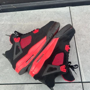 Nike Air Jordan 4 svart/röd sneakers - Nike Air Jordan 4 sneakers i svart och rött med klassisk Jumpman-logga på plösen. Skorna har meshpaneler, snörning och detaljer i plast. Sulan är röd med svarta inslag och insidan är fodrad i rött tyg. Perfekt för dig som gillar streetwear och ikoniska sneakers.