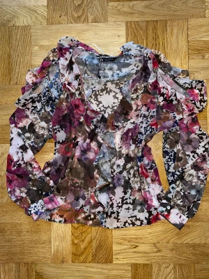 Blommig volangblus från Zara - Säljer en transparent blus från Zara med blommigt mönster i rosa, lila, brun och blå toner. Blusen har långa ärmar, v-ringning och snygga volangdetaljer framtill och längs axlarna. Perfekt för dig som gillar färg och romantiska vibes.