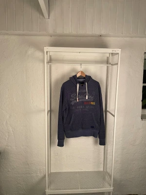 Blå hoodie från Superdry med tryck - Säljer en mörkblå hoodie från Superdry med vintage-tryck och japanska tecken på bröstet. Tröjan har vita snören i huvan, känguruficka och ribbade muddar. Materialet är mjukt och känns som bomull, perfekt för chill dagar. Storlek L.