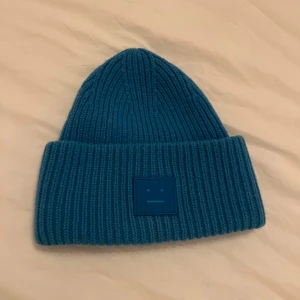  Acne Studios mössa - Snygg blå mössa från Acne Studios med ribbstickad design och uppvikt kant. På framsidan sitter Acne Studios klassiska ansiktslogga i matchande blått. Perfekt för dig som vill ha en stilren och trendig accessoar till kalla dagar.