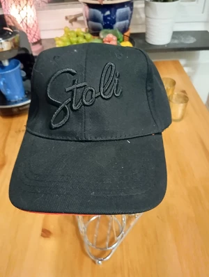 Svart Stoli keps med broderad logga - Snygg svart keps från Stoli med broderad logga framtill. Klassisk form med böjd skärm och diskret röd kant undertill. Perfekt för dig som vill ha en stilren och enkel accessoar. Justerbar baktill för bästa passform.