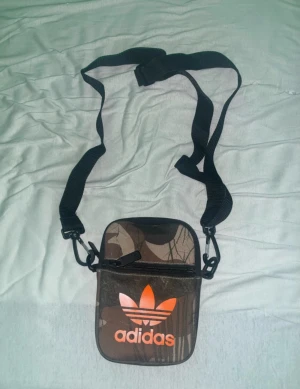 Camouflage axelväska från Adidas - Säljer en liten och smidig axelväska från Adidas med camouflage-mönster och orange logga framtill. Väskan har justerbar svart rem, dragkedja och flera fack inuti. Perfekt för att bära med sig det viktigaste och har en cool streetstil.