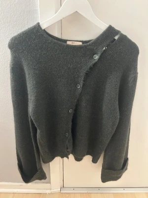 Mörkgrön kofta från Gina Tricot - Mysig mörkgrön kofta från Gina Tricot i stickat material. Koftan har en cool asymmetrisk knappning framtill och långa ärmar med ribbade muddar. Perfekt för lager-på-lager och chill stil under höst och vinter.