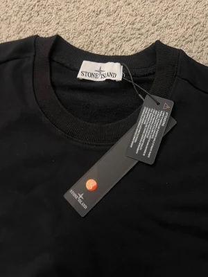 Svart Stone Island sweatshirt - Svart sweatshirt från Stone Island med klassisk rund hals och ribbade muddar. Tröjan har den ikoniska Stone Island-loggan på vänster ärm och är tillverkad i mjuk bomull. Perfekt för dig som gillar stilrena och snygga streetwear-plagg.