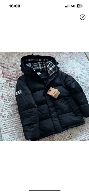 Svart pufferjacka från Burberry - Svart pufferjacka från Burberry med huva och rutig innerfoder i svart, vitt och rött. Jackan har Burberry-logga på ärmen och dragkedja framtill. Perfekt för kalla dagar och har en modern, boxig passform.