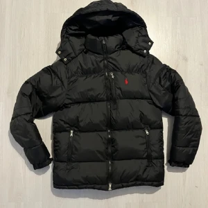 Svart pufferjacka från Polo Ralph Lauren - Svart pufferjacka från Polo Ralph Lauren med röd logga på bröstet. Jackan har huva, flera dragkedjeförsedda fickor och quiltad design. Perfekt för kalla dagar med sin tjocka och fluffiga look. Snygg och stilren, passar dig som gillar streetwear.Det ska stå polo på dragkedjan. Den har några små hål men det är inget stort man kan även tejpa allt möjligt funkar. 