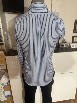 Ralph Lauren skjorta - Snygg skjorta från Polo Ralph Lauren med klassiska svarta ränder på vit botten. Skjortan har lång ärm, knappar framtill och en stilren krage. Materialet är mjuk bomull och loggan är broderad på bröstet. Perfekt för dig som gillar en clean och tidlös look.