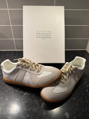 Maison Margiela Replica sneakers vit/beige - Maison Margiela Replica sneakers i vit och beige med klassisk gummisula. Ovandel i mjukt skinn och mocka, med rund tå och snörning. Snygg retrostil och neutrala färger som passar till det mesta. Ikonisk design med Margielas signatur på sulan och innersulan.
