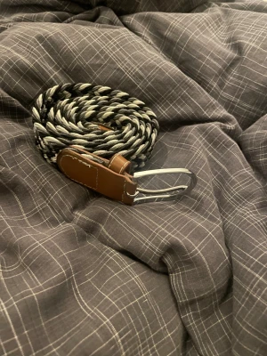 Flätat bälte   - Snyggt flätat bälte i svart och vitt med bruna skinn-detaljer vid spännet. Bältet har ett silvrigt metallspänne och är gjort i ett mjukt, flexibelt material som gör det bekvämt att bära. Perfekt accessoar för att lyfta din outfit med en cool kontrast.