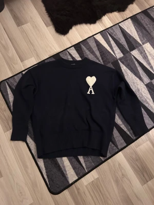 Mörkblå stickad tröja från Ami - Snygg mörkblå stickad tröja från Ami med ett vitt hjärta och bokstaven A broderat på bröstet. Tröjan har rund hals, ribbade muddar och lång ärm. Perfekt för dig som gillar stilrena detaljer och vill ha något som sticker ut lite.