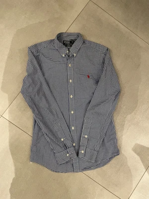 Ralph Lauren skjorta - Tja säljer en snygg Slim fit skjorta från Ralph Lauren i blått och vitt rutigt mönster. Sjukt snygg med krage, långa ärmar och vita knappar framtill. Broderad röd Polo-logga på bröstet. Skjortan är gjord i mjuk bomull och har en clean, modern look. Passar perfekt till ett kalas eller fest eller bara till vardagen!🤩perfekt skick ungefär som nytt storleken är S passar dig som är runt 175-180cm lång skulle jag tro!! Har du övriga frågor hör av dig!🙌🏼mvh EM_resell
