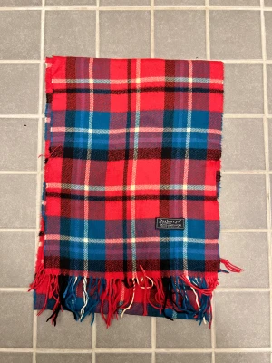 Rutig röd och blå halsduk från Burberry - Klassisk rutig halsduk från Burberrys i 100% ull. Halsduken har fransar i ändarna och den ikoniska Burberrys-etiketten. Perfekt accessoar för att lyfta din outfit. Anledningen att de stör ”burberrys” är för att de är en äldre modell.