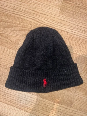Svart stickad mössa från Polo Ralph Lauren - Svart ribbstickad mössa från Polo Ralph Lauren med klassisk röd logobroderi framtill. Tillverkad i 100% bomull och har en uppvikt kant för extra stilpoäng. Perfekt för dig som vill ha en clean look med en touch av lyx.