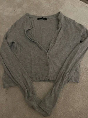 Grå kofta från 157, croppad XS - Grå croppad kofta från 157 i storlek XS. Tröjan har lång ärm, ribbade muddar och knappar framtill. Perfekt att slänga över en topp för en chill look. Materialet är mjukt och känns som bomull eller syntet. Enkel och stilren design.