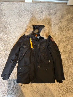 Svart Parajumpers parkajacka med päls - Svart parkajacka från Parajumpers med äkta päls runt huvan, flera fickor med dragkedja och knappar, samt den ikoniska gula remmen. Jackan har lång ärm och detaljerad patch på axeln. Perfekt för kalla dagar med snygg och funktionell design.