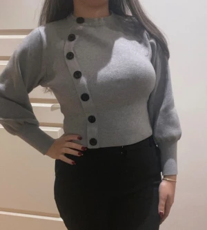 Grå tröja med svarta knappar - Snygg grå långärmad tröja med svarta knappar längs sidan framtill. Modellen är croppad med långärmade puffärmar och ribbade muddar. Materialet känns mjukt mot huden. Perfekt för dig som gillar stilrena detaljer och vill ha något som sticker ut. 