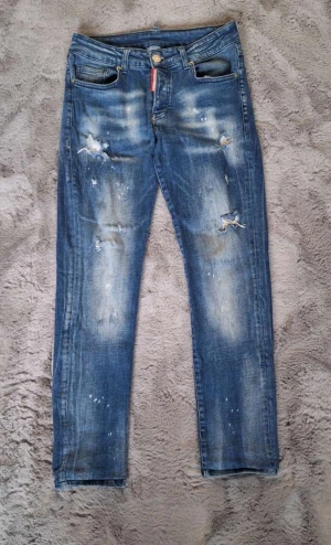 Dsquared2 ICON blå jeans med slitningar - Säljer ett par Dsquared2 ICON jeans i blå tvätt med coola slitningar och färgstänk. Klassisk femficksmodell med röd Dsquared2-tag vid fickan. Stl 44, bra skick. 