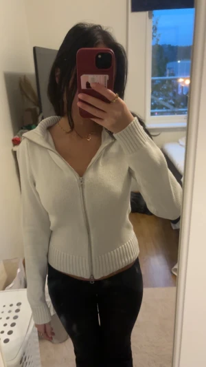 Vit stickad hoodie med dragkedja - Säljer en vit stickad hoodie med huva och hel dragkedja framtill. Tröjan har ribbade muddar vid ärmslut och nederkant, samt en snygg normal passform. Perfekt för dig som gillar stilrena och bekväma plagg med lite edge.