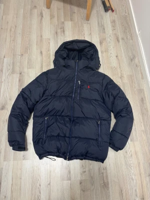 Ralph lauren puffer jacka - Riktig snygg mörkblå pufferjacka från Polo Ralph Lauren med huva och dragkedja framtill. Jackan har en röd Polo-logga på bröstet, snörning vid nederkant och två sidofickor med dragkedja. Perfekt för kalla dagar och har en riktigt schysst, puffig look. För mer info eller bilder så är det bara att skriva.