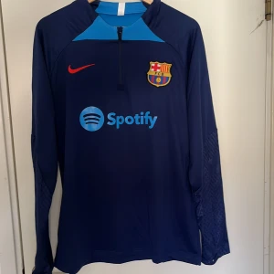 Blå långärmad FC Barcelona Nike-tröja - Säljer en långärmad FC Barcelona träningströja från Nike i mörkblått med ljusblå detaljer. Tröjan har halv dragkedja, klubbmärke på bröstet, röd Nike-logga och Spotify som sponsor. Tillverkad i lätt och funktionellt material, perfekt för fotbollsträning. Storlek L men passar även M!    Pris kan diskuteras + skriv till mig vid eventuella frågor🤩