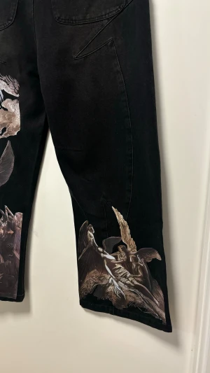 Svarta jeans med ängeltryck baggy jeans  - Unika svarta jeans med croppad benlängd och bred passform. Framsidan har detaljerade tryck med änglar, moln och strålar i beige och grå toner. Snygga sömmar och stora bakfickor ger extra edge. Perfekt för dig som vill sticka ut med konstnärlig vibe.