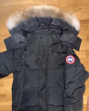 Svart dunjacka från Canada Goose - Säljer en svart dunjacka från Canada Goose med ikonisk röd och vit logga på ärmen. Jackan har en stor huva med fluffig päls, knappar och dragkedja framtill. Perfekt för kalla vinterdagar och levereras i originalväska.