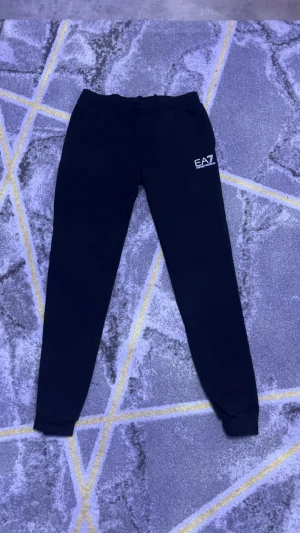 Svarta mjukisbyxor EA7 Emporio Armani - Svarta mjukisbyxor från EA7 Emporio Armani med logga tryckt på vänster lår. Byxorna har elastisk midja med snörning och ribbade muddar vid bensluten. Perfekt för chill eller träning, stilren och enkel design.