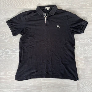 Svart Burberry pikétröja - Svart pikétröja från Burberry Brit i 100% bomull. Klassisk krage, korta ärmar och knäppning med Burberrys ikoniska rutiga tyg på insidan. Liten broderad logga på bröstet.