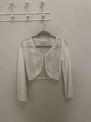 Vit spetsig kofta från H&M - Säljer en vit kofta från H&M med lång ärm och croppad passform. Den är barnstorlek i storleken 146/152!!! Toppen är gjord i spets med ett snyggt mönster och har en öppen front med rundad kant. Knäpps med i knapp i mitten. Perfekt att styla över linne eller klänning för en trendig look.