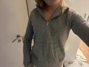 Grå hoodie med dragkedja - Mysig grå hoodie med dragkedja framtill och huva med snören. Tröjan har två stora fickor på magen och ribbade muddar vid ärmslut och nederkant. Perfekt för chill dagar eller när du vill ha något bekvämt på dig. 