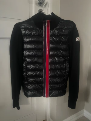 Svart Moncler pufferjacka med stickade ärmar - Svart pufferjacka från Moncler med glansig quiltad front och stickade ärmar. Röd och blå randig dragkedja framtill, samt Moncler-logga på vänster ärm. Jackan har hög krage och långärmad passform, perfekt för dig som gillar stilren design med sportig touch. Nykligen köpt på Vinted, men tyvärr blev inte storleken som jag trodde, passar mer 158-164cm enl min mening. Jag artikelverifierade den, så den är äkta. Endast provad här och enl förra ägaren i nyskick och det är den verkligen! Storlek 14A