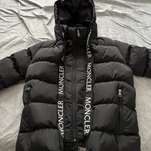 Svart dunjacka från Moncler med huva - Säljer en svart dunjacka från Moncler med stor huva och tydliga logotexter längs dragkedjan. Jackan har två fickor med dragkedja och klassisk Moncler-logga på ärmen. Perfekt för kalla dagar och riktigt snygg streetstil.