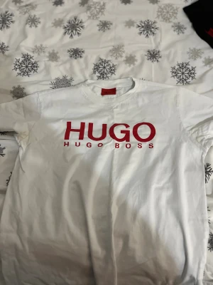 Vit HUGO BOSS t-shirt med röd logga - Snygg vit t-shirt från HUGO BOSS med stor röd HUGO-logga på bröstet. Klassisk rund hals och korta ärmar. Tillverkad i mjuk bomull som känns skön mot huden. Perfekt för dig som gillar stilrena och trendiga plagg med tydlig branding.