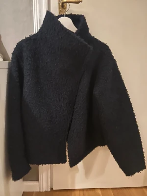 mörkblå teddyjacka med hög krage - Mysig mörkblå teddyjacka med hög krage och asymmetrisk front. Jackan har en fluffig, strukturerad yta och stängs med tryckknappar. Perfekt för dig som gillar en chill och trendig look under kyliga dagar. Knapparna är lite trasiga, har kvar extra knappen men inte sytt på den. Den översta knappen är bara att ni in lite mer 