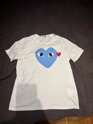 Vit Comme des Garçons Play t-shirt - Vit t-shirt från Comme des Garçons Play med stort blått hjärta med ögon tryckt på bröstet och ett mindre rött hjärta broderat bredvid. Klassisk rund hals och korta ärmar. Tillverkad i mjuk bomull för skön känsla.