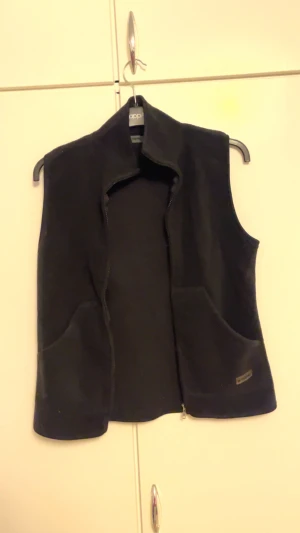 Svart fleeceväst från B-Young Essential - Svart fleeceväst från B-Young Essential med hög krage och två stora fickor framtill. Västen har dragkedja och är perfekt att slänga över en hoodie eller långärmad tröja. Enkel och clean design som funkar till många olika stilar.