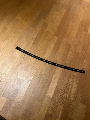 Svart textbälte från VOX - Svart bälte från VOX med vit text och grafiska detaljer längs hela bandet. Bältet har en stilren, rektangulär spänne i svart och är tillverkat i slitstark syntet. Perfekt för att ge din outfit en cool och modern touch.