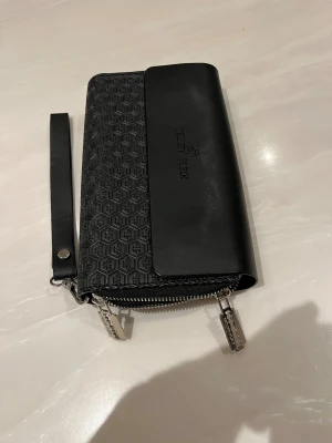 Svart plånbok från Philipp Plein - Snygg svart plånbok från Philipp Plein med präglat hexagonmönster och logga på framsidan. Plånboken har silverfärgade dragkedjor, flera fack för kort och kontanter samt en avtagbar handledsrem. Tillverkad i skinn med coola detaljer och stilren design.