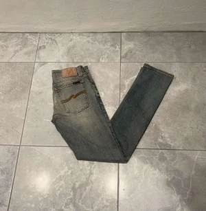 Nudie Jeans blå straight fit - Snygga blå jeans från Nudie Jeans med ljusbrun broderad detalj på bakfickan. Jeansen har en rak passform och är tillverkade i slitstarkt denimtyg. Perfekta för dig som gillar en enkel look. Storleken är W26 L32