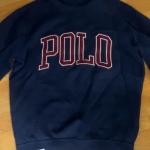 Mörkblå sweatshirt från Polo Ralph Lauren - Mörkblå sweatshirt från Polo Ralph Lauren med stor POLO-logga i vitt och rött framtill. Klassisk crewneck-modell med långa ärmar och liten röd broderad logga på ärmen. Tillverkad i mjuk bomull, perfekt för en chill och stilren look.