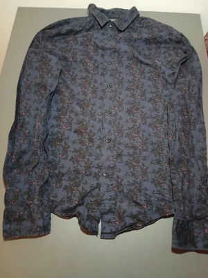 Blommig mörkblå skjorta med knappar - Snygg mörkblå skjorta från Zara Man med blommigt mönster i svart och diskreta rosa detaljer. Skjortan har klassisk krage, långa ärmar och knäppning framtill. Perfekt för dig som vill sticka ut med en cool och unik stil.