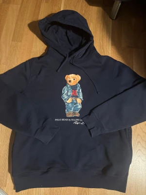 Mörkblå hoodie Polo Bear Ralph Lauren - Snygg mörkblå hoodie från Polo Ralph Lauren med ikoniskt Polo Bear-tryck på bröstet. Tröjan har huva med snörning och långa ärmar. Materialet är mjukt och skönt, perfekt för chill dagar. Klassisk streetwear-vibe med coolt motiv framtill.