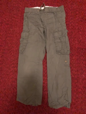 Gröna cargopants från L.O.G.G. strl 134 - Säljer ett par gröna cargopants från L.O.G.G. i storlek 134. Byxorna har flera praktiska fickor på sidorna, elastisk midja med snörning och är tillverkade i mjuk bomull. Perfekt för dig som gillar avslappnad stil och funktionella detaljer.