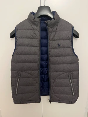 Vändbar väst från Polo Ralph Lauren - Snygg och quiltad väst från Polo Ralph Lauren i grått och marinblått. Västen är vändbar med dragkedja, två fickor med zip och hög krage. Den är i superbra skick. Skriv gärna vid funderingar, priset är diskuterbart!
