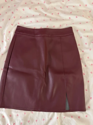 Vinröd skinnkjol från Bik Bok - Vinröd kort skinnkjol från Bik Bok i storlek 34. Kjolen har en stilren design med en liten slits framtill och dragkedja bak. Snygg och tight passform.