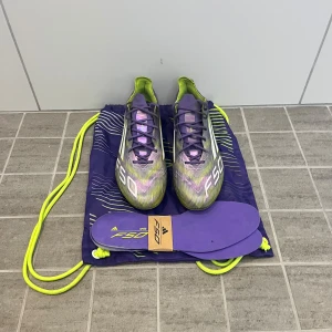 Adidas F50 elit - Säljer ett par Adidas F50 fotbollsskor i en snygg lila och limegrön färg med vita detaljer och snörning. Skorna har en låg profil, platt sula med dobbar och är tillverkade i syntetmaterial för lätthet och snabbhet. Medföljer matchande lila innersulor och väska.