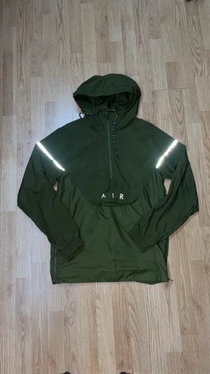 Nike Air grön vindjacka med huva - Snygg grön Nike Air vindjacka med reflekterande detaljer på ärmarna och tryck på ryggen. Jackan har huva med snörning, half zip framtill och elastiska muddar vid ärmslut. Perfekt för dig som gillar streetwear och sportig stil. Obs lite av reflexen har gårt bort på båda armarna